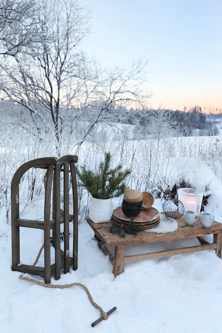 Crockery on rustic wooden table, Christmas decorations and wooden sledge in snowy landscape