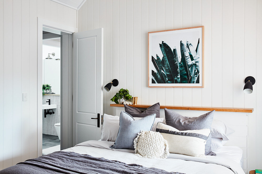 Scatter cushions on double bed against narrow shelf in bedroom with white wood-clad walls