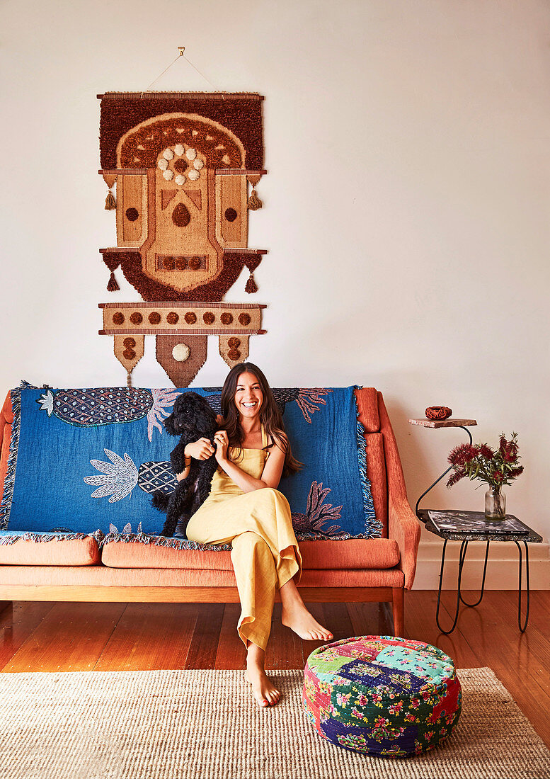 Junge Frau mit Hund sitzt auf Sofa mit Decke, darüber Ethno-Wandbehang
