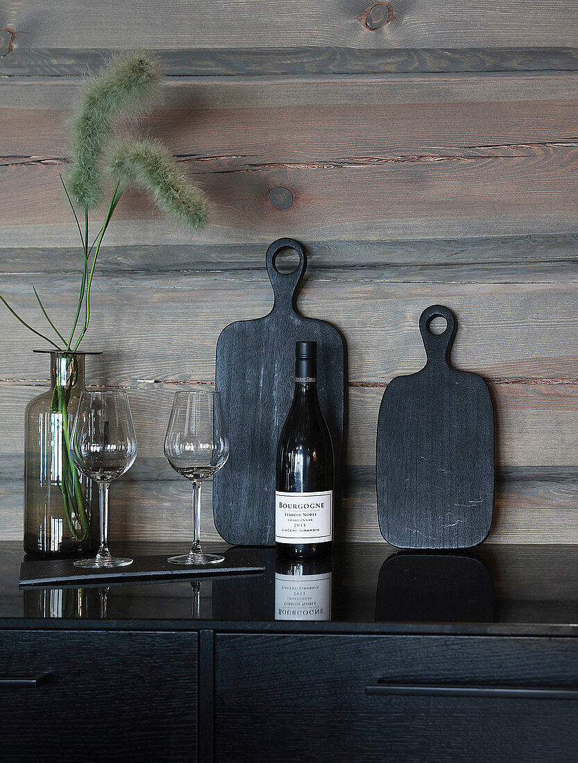 Black chopping boards, bottle of wine and glasses against wooden wall