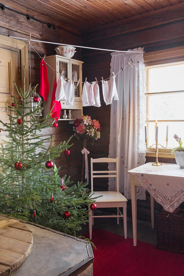Christmas tree decorated with red baubles, chair and table in rustic room with wood-clad walls and laundry hung from washing line
