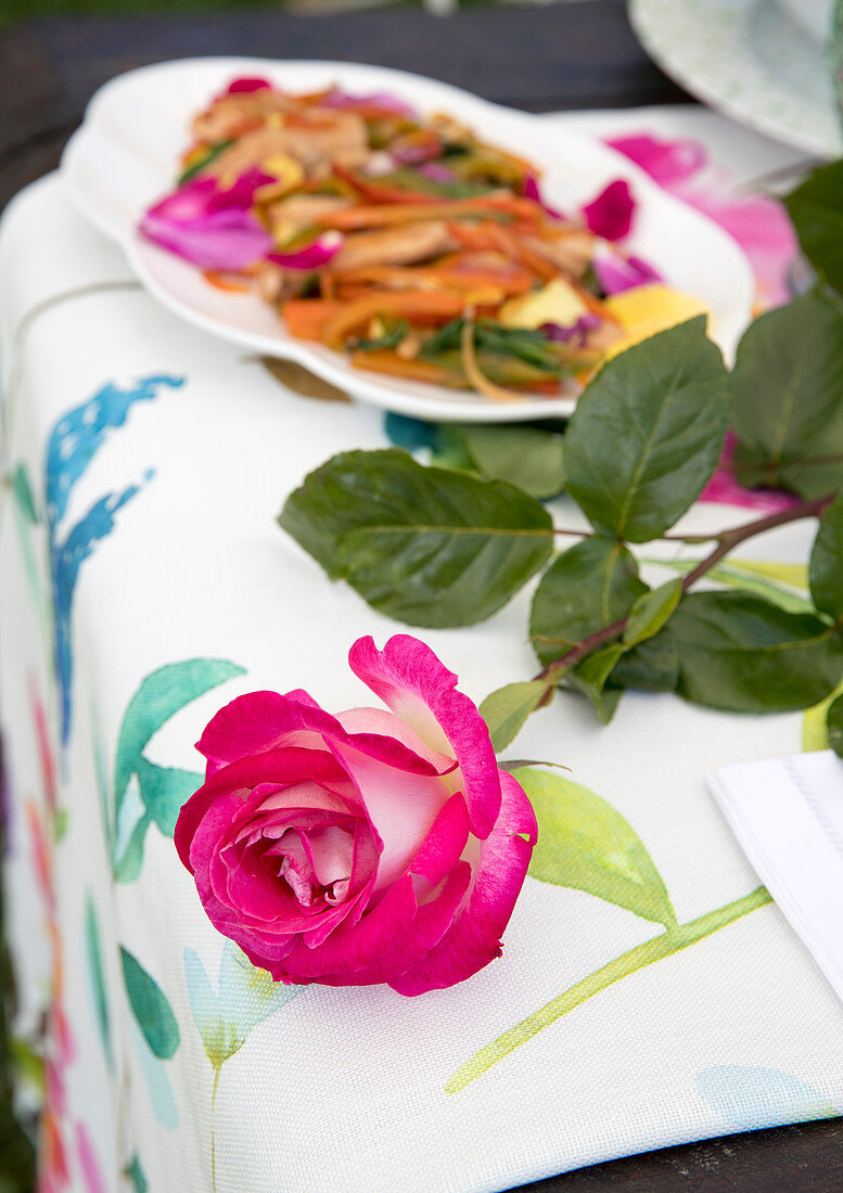 Pinkfarbene Rose und sautiertes Hähnchen mit Peperoni und Rosenblättern auf Gartentisch