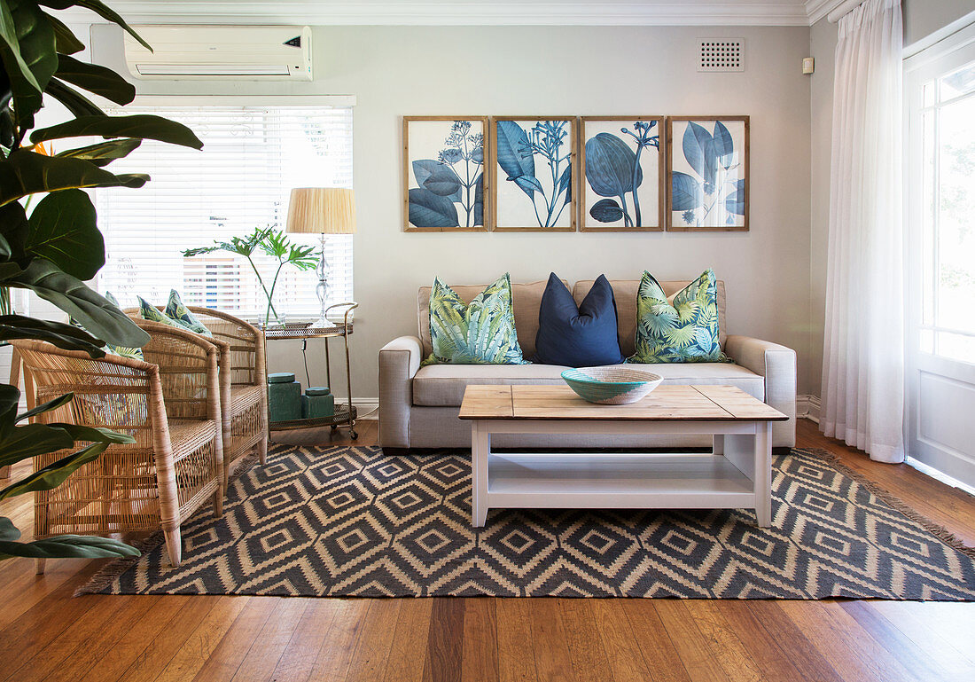 Rattansessel, Couch und Couchtisch auf Teppich mit geometrischem Muster und botanische Drucke an der Wand im Wohnzimmer
