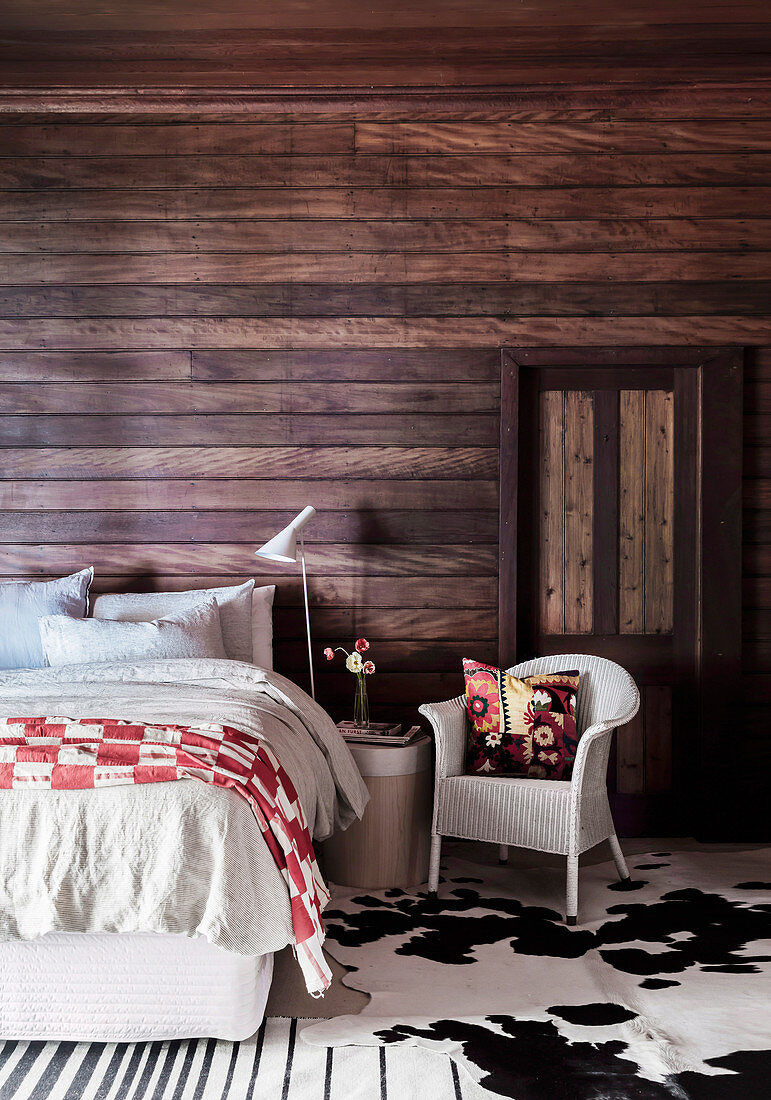 Doppelbett, Nachttisch und Rattansessel im Schlafzimmer mit dunkler Holzverkleidung