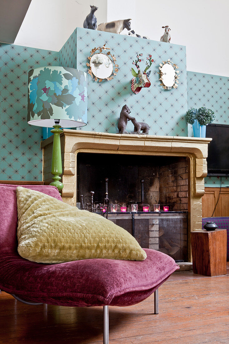 Velvet easy chair with large cushion in front of old fireplace and patterned wallpaper