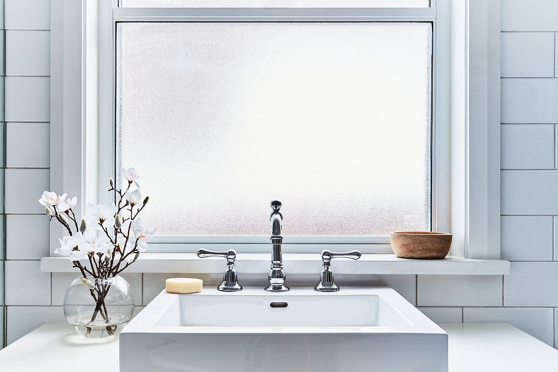 Weißer Waschtisch mit Aufsatzbecken und Kugelvase mit Magnolienblüten vor Fenster im Badezimmer