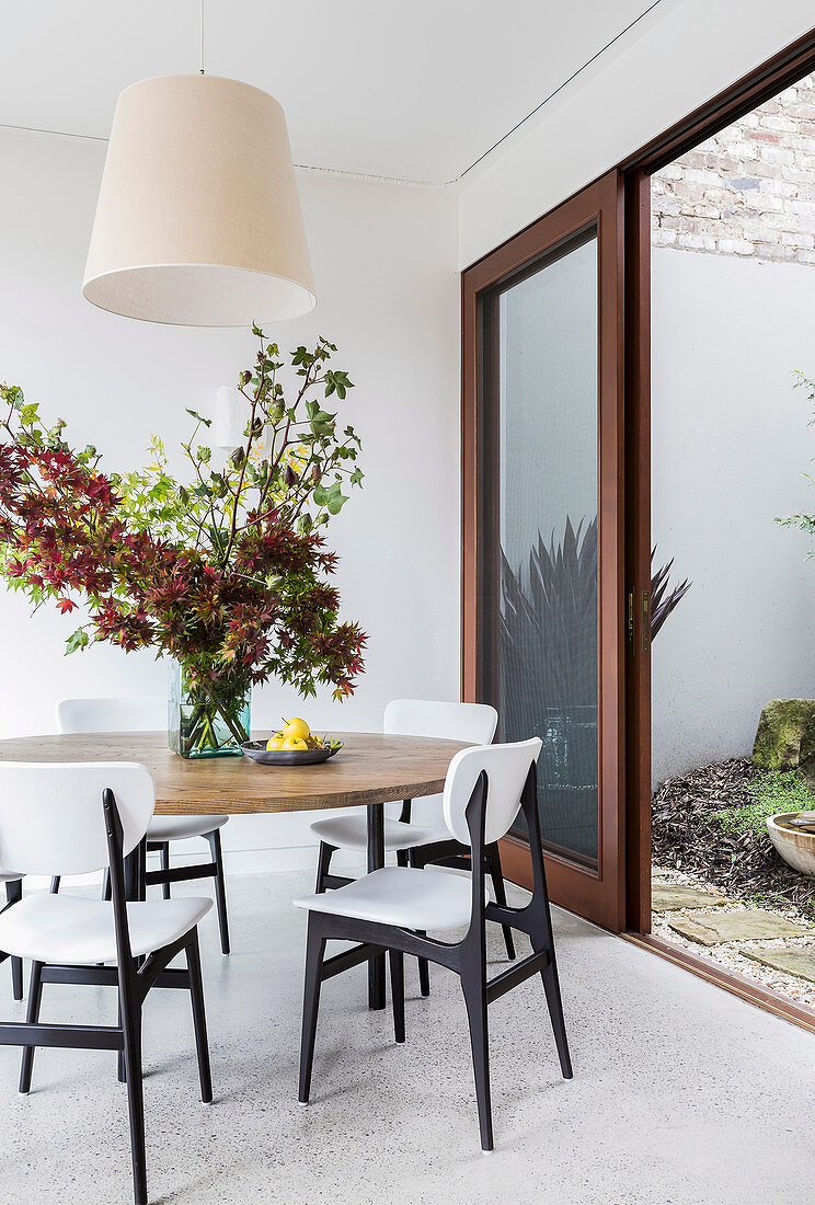 Runder Tisch mit Blätterzweigen und weiße Stühle vor offener Terrassenschiebetür