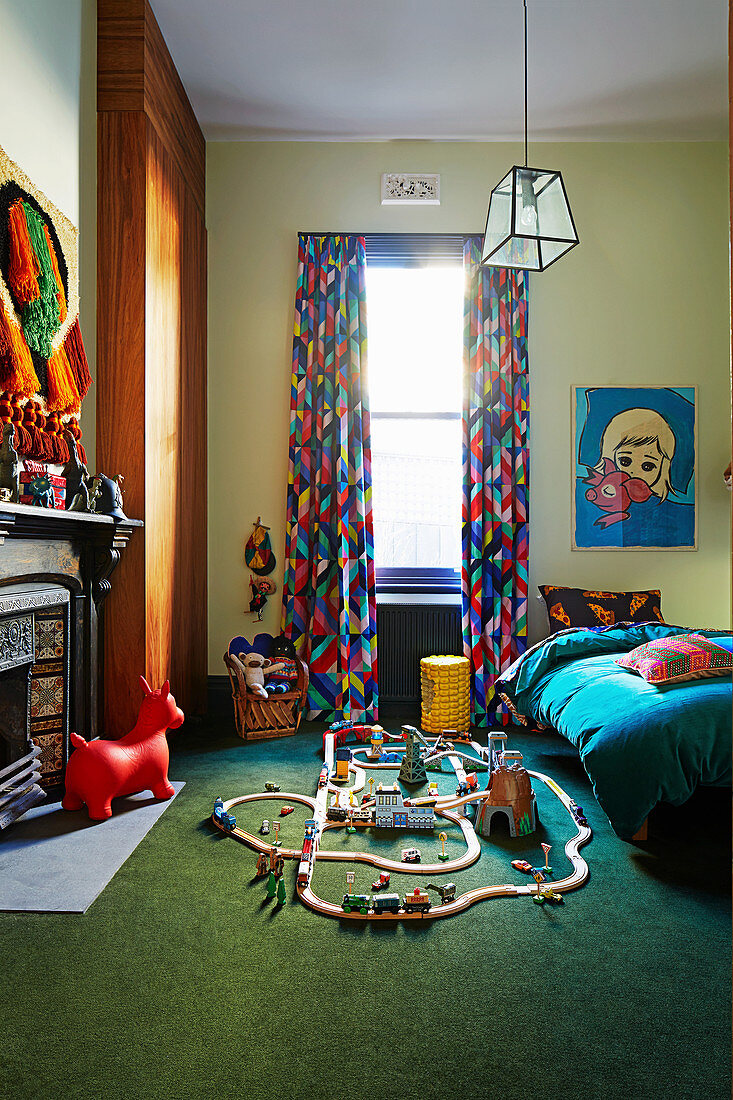 Holzeisenbahn auf grünem Teppichboden, Bett, bunter Vorhang und Kamin im Kinderzimmer