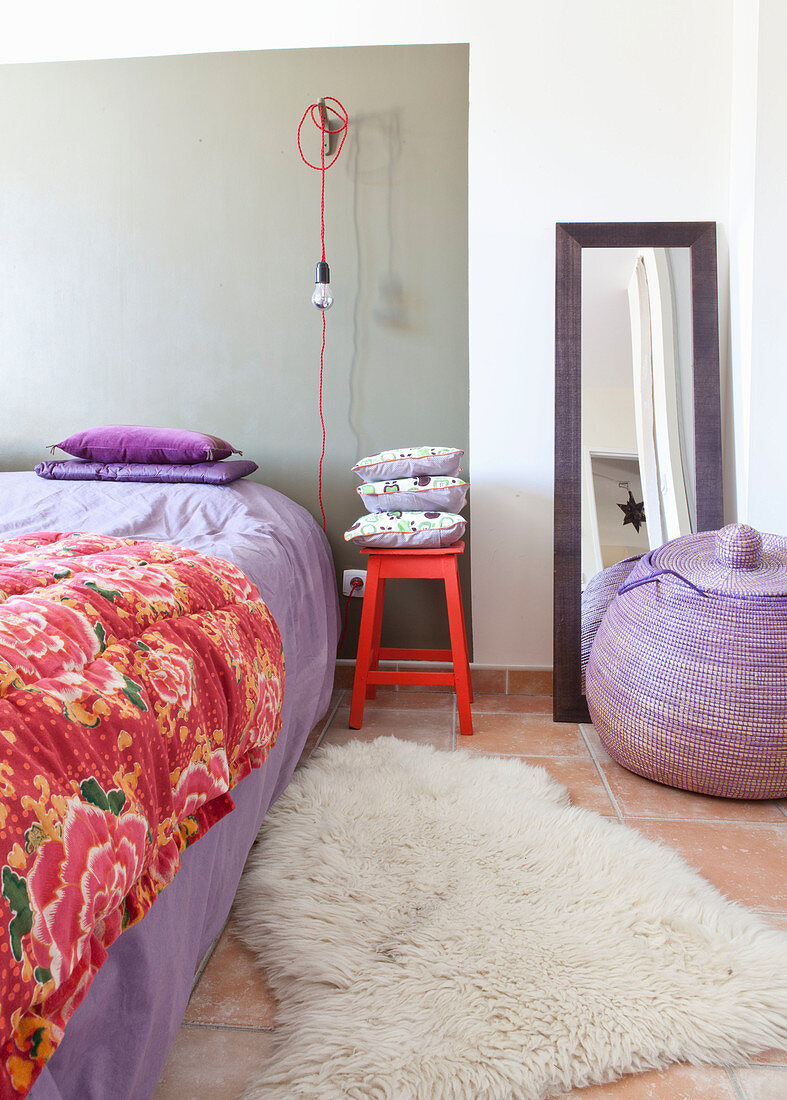 Bett mit lila Bettwäsche, roter Hocker mit Kissen und Glübirne mit rotem Kabel, lila Korb vor Standspiegel im Schlafzimmer