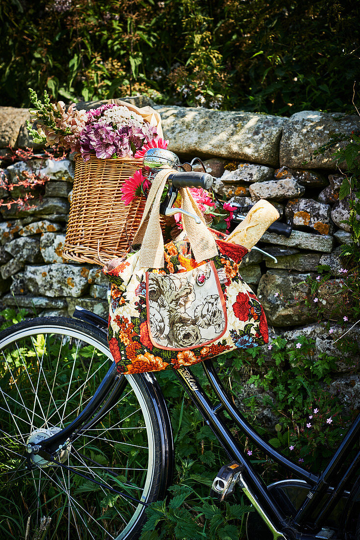 Weidenkorb mit Blumen und blumige Einkaufstasche mit Baguette am Fahrrad