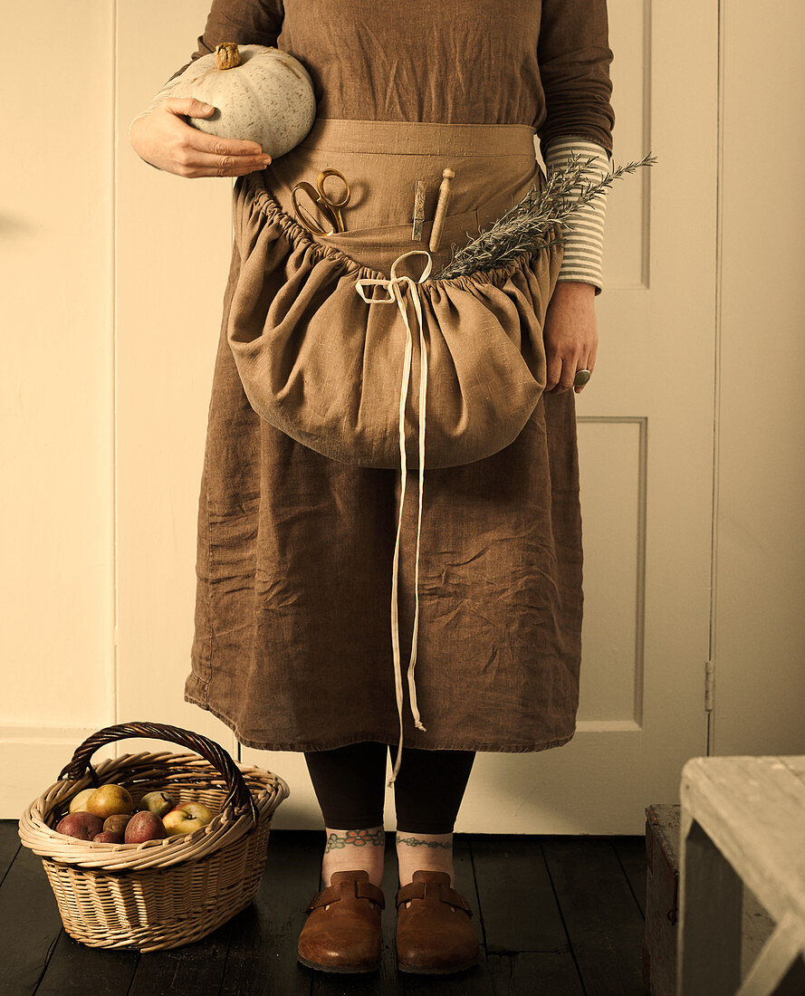 Woman wearing brown clothing holding pumpkin and with basket of freshly picked apples
