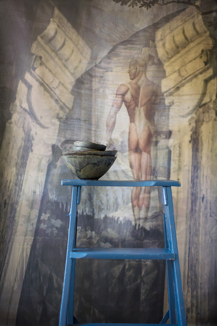 Stapel dunkler Schälchen auf einer blauen Leiter vor einem Wandbild