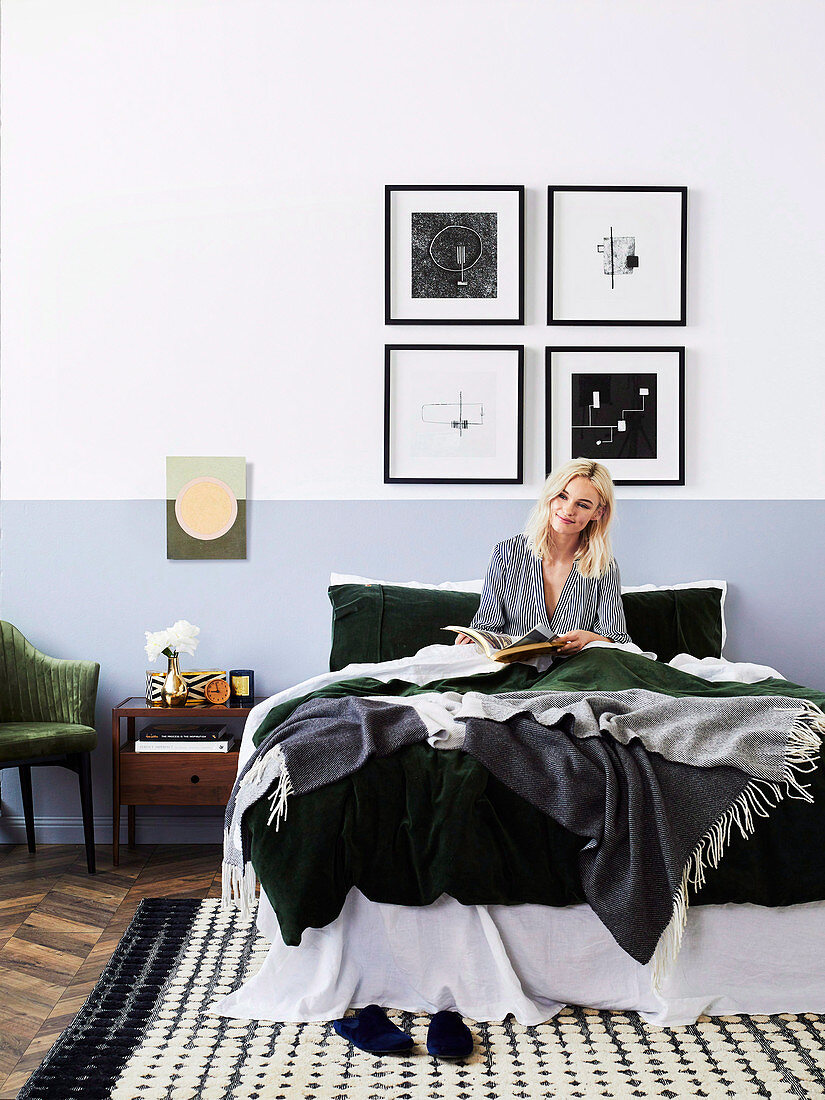 Junge Frau mit Buch im Bett, gerahmte Kunstwerke an der Wand