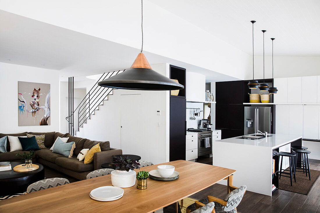 Lange Esstisch vor Lounge mit Polstersofa, im Hintergrund schwarz-weiße Küche mit Theke in offenem Wohnraum