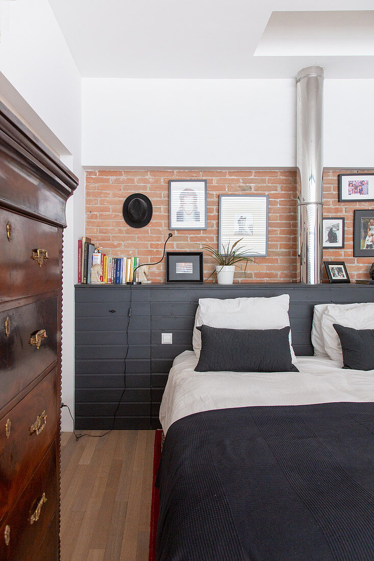 Double bed against black sideboard and brick wall in bedroom with antique chest of drawers in foreground