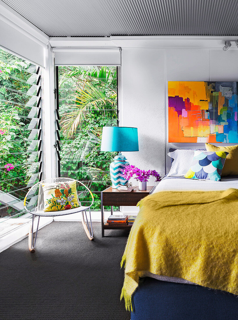Doppelbett mit senfgelber Tagesdecke, Nachttisch mit blauer Tischlampe und Schaukelstuhl im Schlafzimmer, buntes Kunstwerk an der Wand