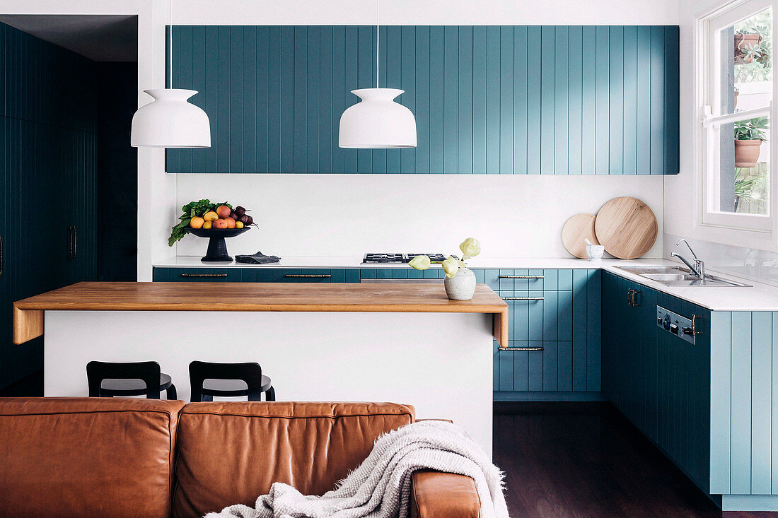 Blick über Ledersofa auf Einbauküche mit blau-grau lackierter Holzfront und Kücheninsel