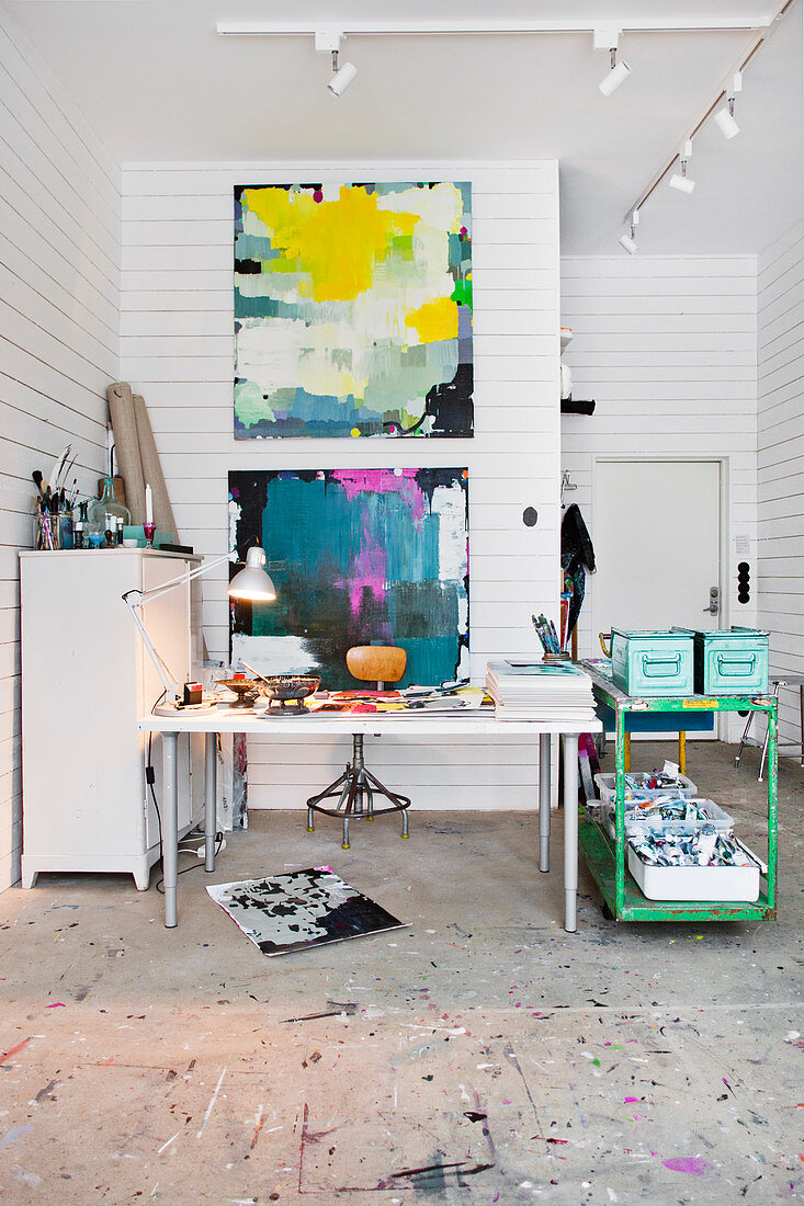 Desk and green, vintage serving trolley in artist's studio with paintings on white wooden wall