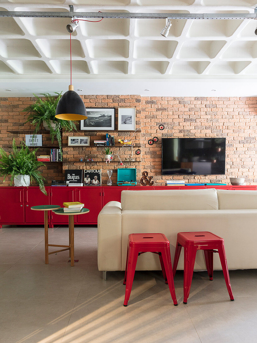 Rote Hocker am Sofa, im Hintergrund rote Unterschränke an Backsteinwand in offenem Wohnraum