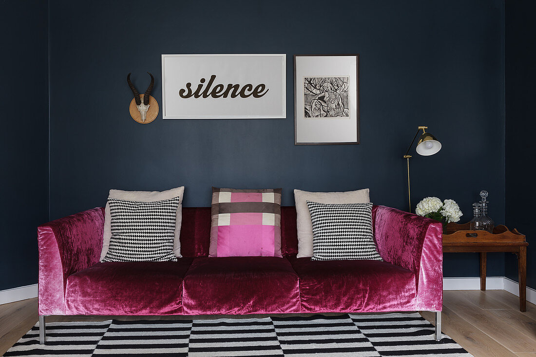 Couch mit pinkfarbenem Samtbezug und Kissen auf schwarz-weiß gestreiftem Teppich vor dunkler Wand