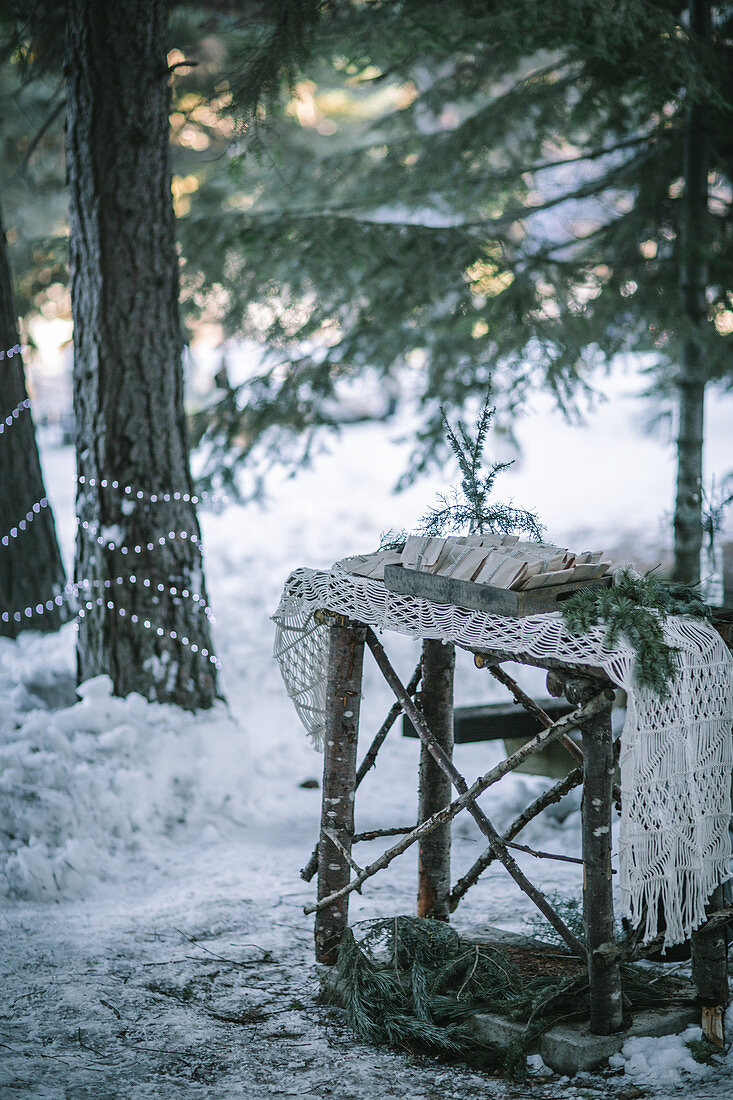 Kleiner Holztisch mit Makramee-Tischläufer und Geschenken im verschneiten Wald