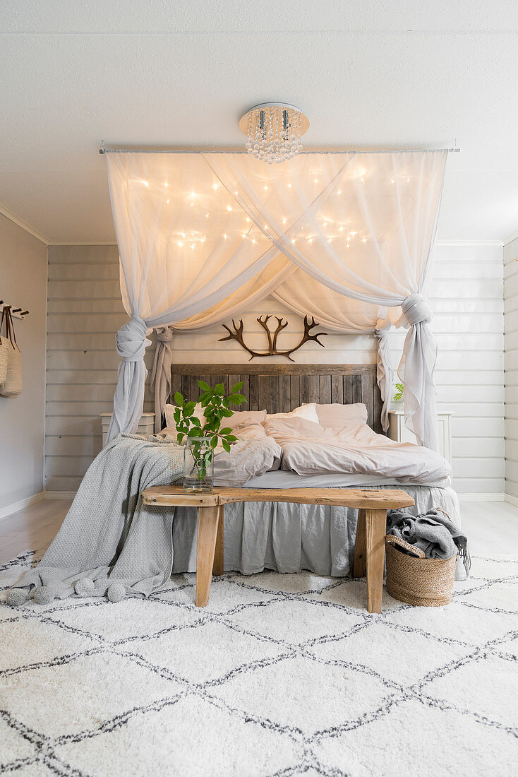 Double bed below canopy with lights and wooden bench in pale bedroom