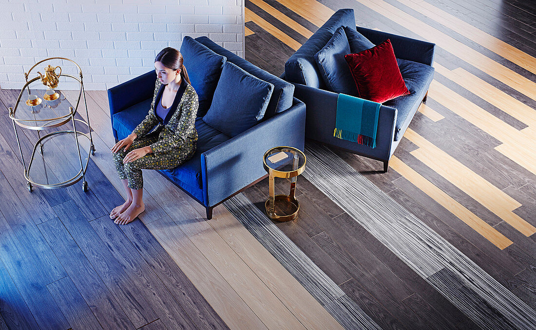 Wohnraum mit Holzboden in verschiedenen Schattierungen, Frau sitzt auf blauem Zweisitzer, daneben Servierwagen und Beistelltisch