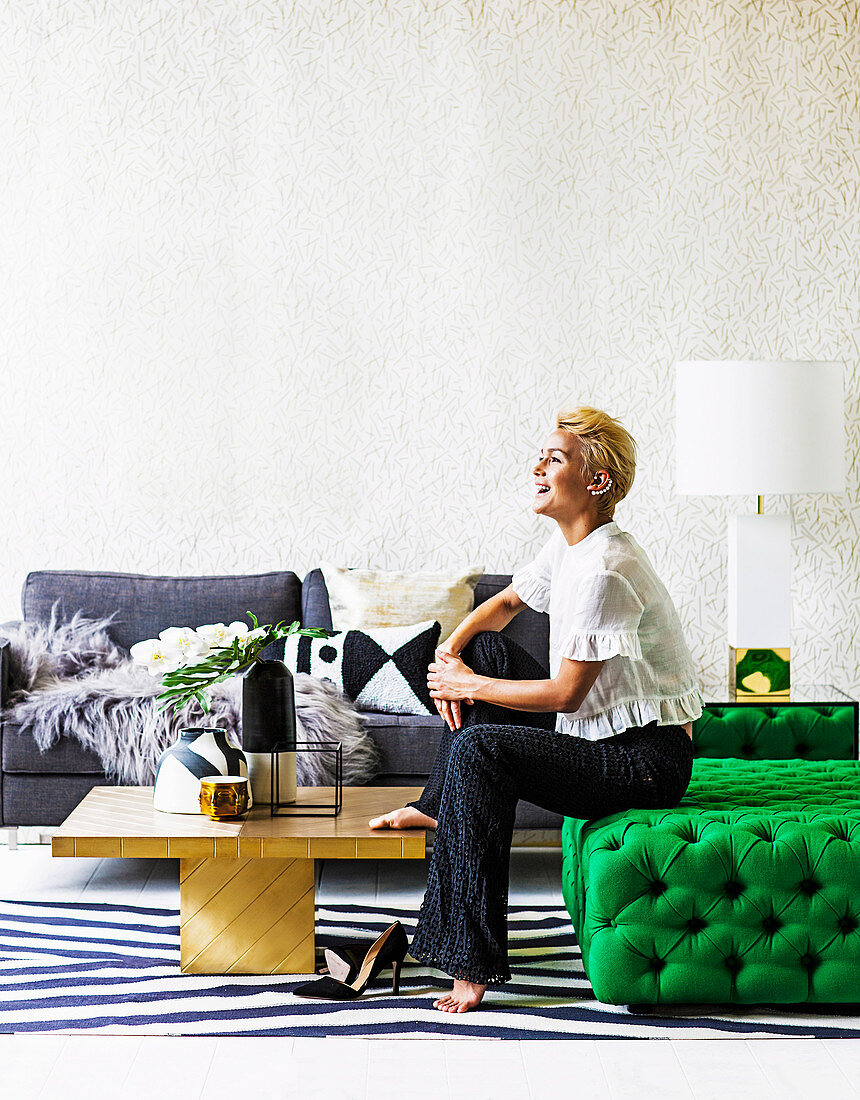 Blonde Frau sitzt auf grünem Ottoman, goldfarbener Couchtisch und graues Sofa im Wohnzimmer