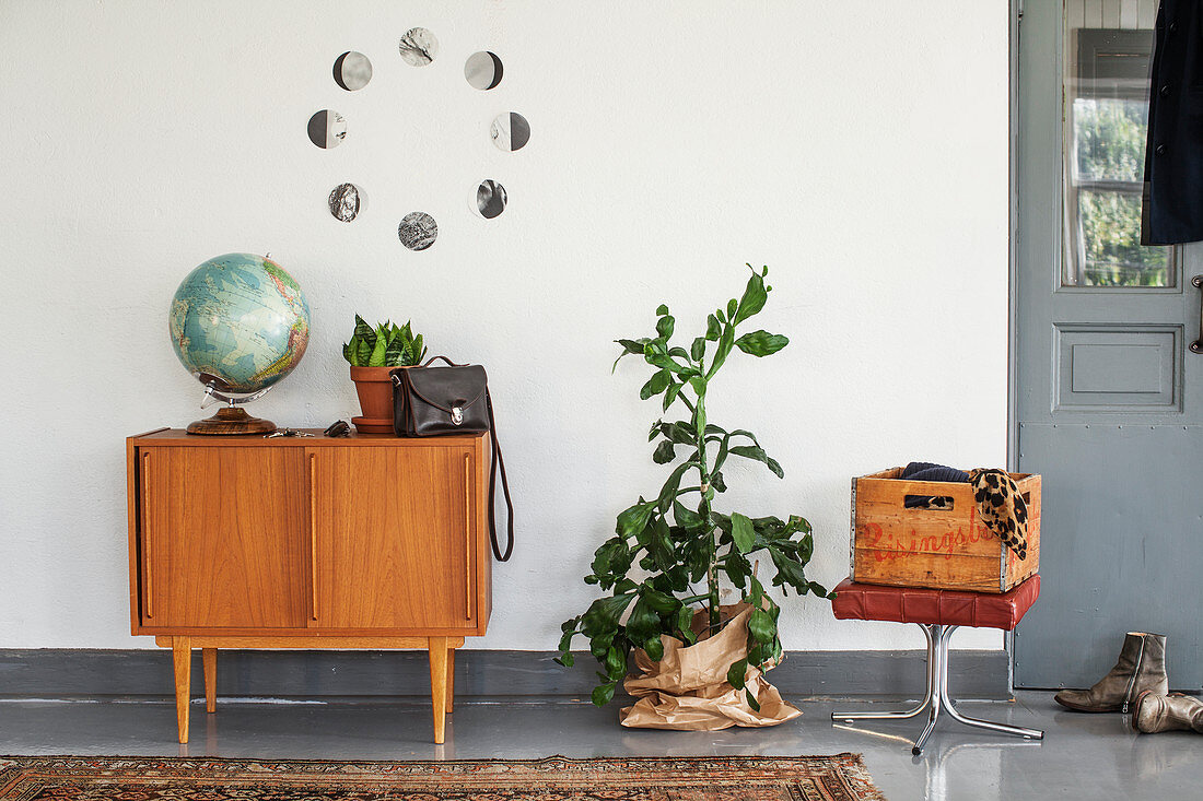 Globe on sideboard below circular arrangement of black-and-white photos on wall, houseplants and wooden crate on stool