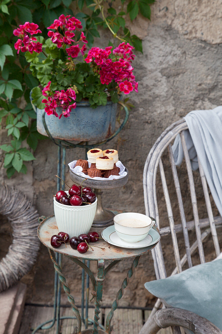 Schälchen mit Kirschen, Kaffeetasse und Etagere mit Gebäck auf Vintage Tischchen auf der Terrasse
