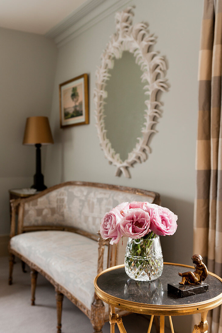Pink roses in glass vase on round occasional table, in the background is an antique settee beneath a plaster framed mirror