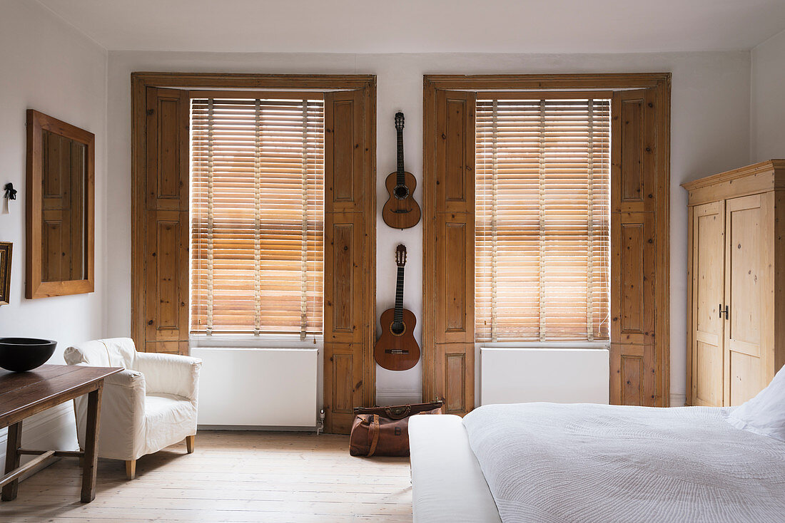 Classical guitars hung up on wall of bedroom with pine window shutters and white armchair