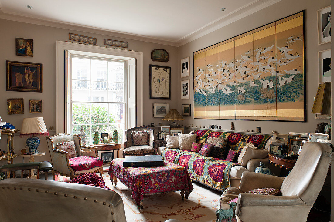 Eclectically furnished living room with assorted artworks, cream leather and wood chairs in the foreground, the sofa is covered with a matrimonial textile, above the sofa hangs a Japanese screen with birds