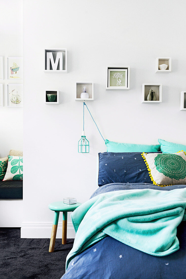 Bed with bedclothes in aqua tones, shelving set above, pendant lamp and stool as bedside table
