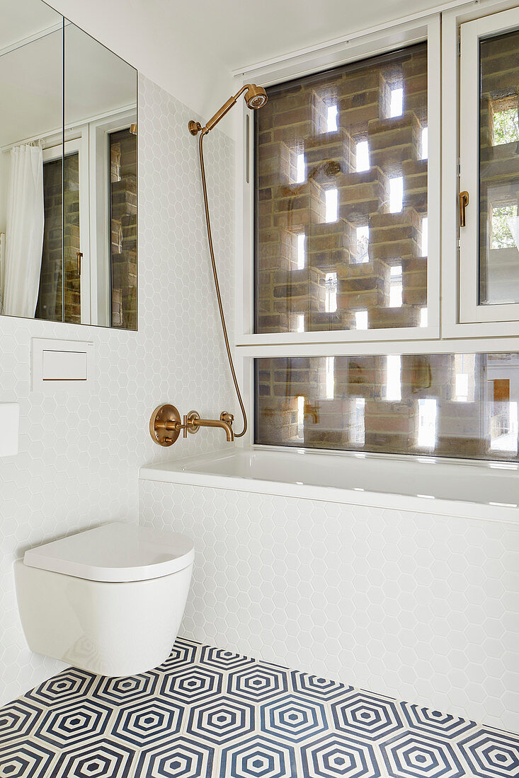 Shower head over bathtub, mirror, and toilet in bathroom in bathroom with window behind perforated brick façade
