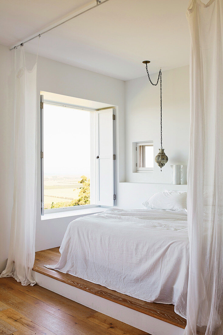 Doppelbett mit weißer Decke auf Podest im Schlafbereich mit weißem Vorhang