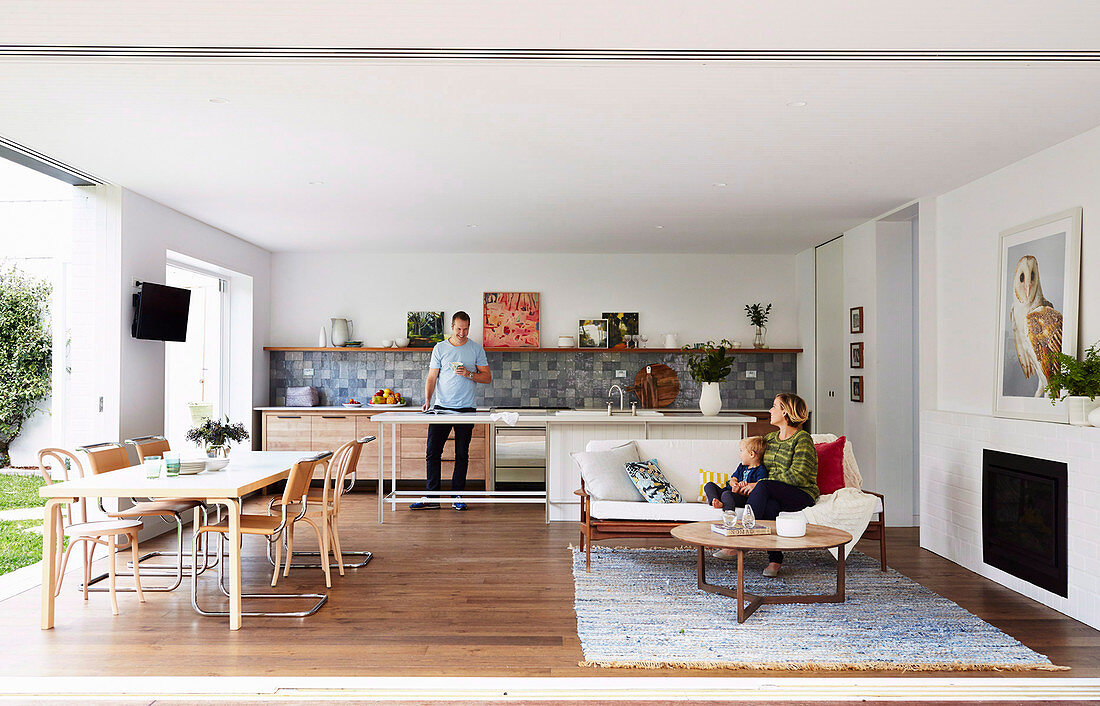 Familie in offenem Wohnraum mit Küche, Sofa vor Kamin und Essbereich vor geöffneten Terrassentüren