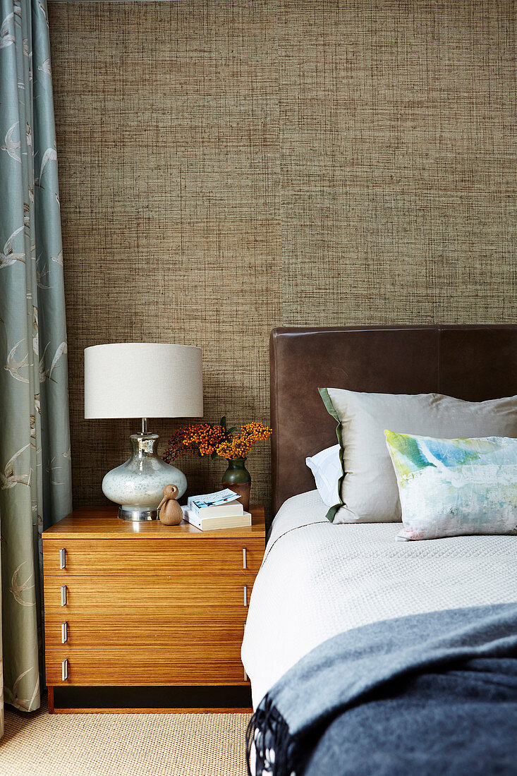 Double bed with leather headboard, bedside table with lamp and textile wallpaper in the bedroom