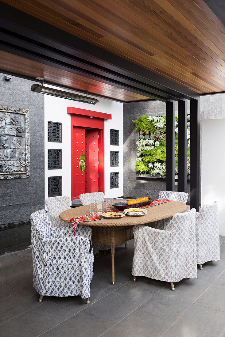 Überdachte Terrasse mit Rattantisch, Hussenstühlen, vertikaler Bepflanung und roter, chinesischer Tür