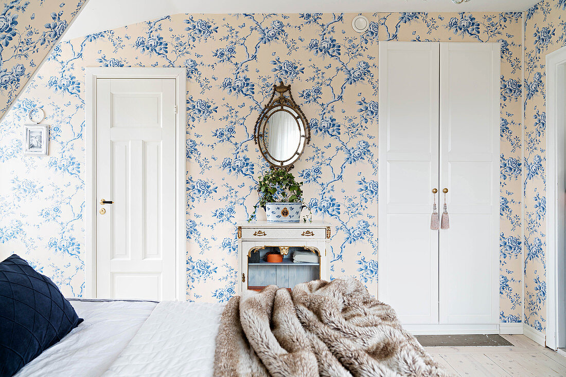 Blick über Doppelbett auf Wandtisch zwischen Tür und Einbauschrank in romantischem Schlafzimmer mit floraler Tapete
