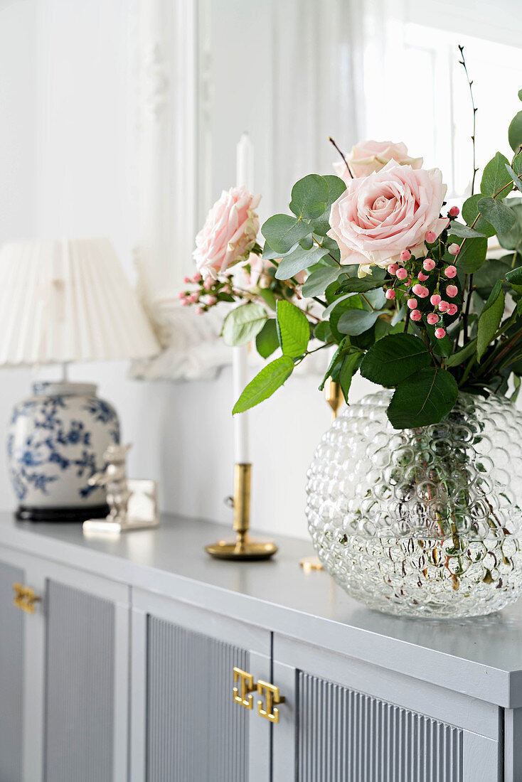 Bouquet of roses in glass vase, candlestick and table lamp on pale grey sideboard