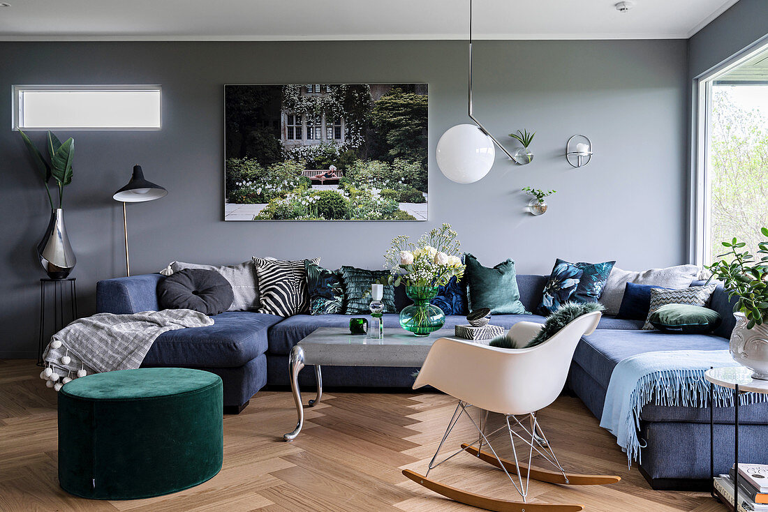 Blaues Loungesofa, grüner Sitzpouf und Klassiker-Schaukelstuhl im Wohnzimmer mit grauer Wand