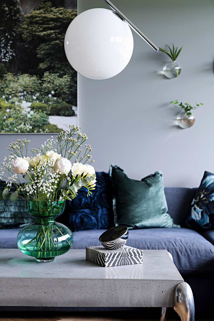 Vase of flowers on coffee table with concrete top below spherical lamp and in front of sofa with scatter cushions in living room with grey wall