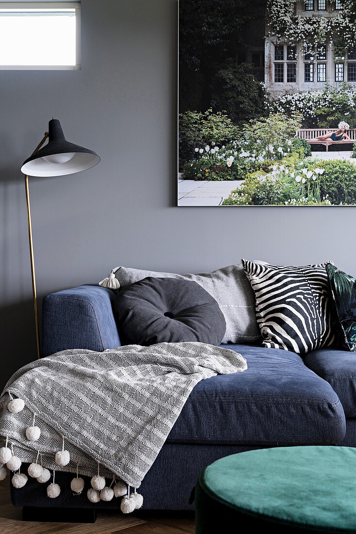 Blaues Loungesofa mit Kissen und Plaid, Stehlampe und Bild an grauer Wand im Wohnzimmer
