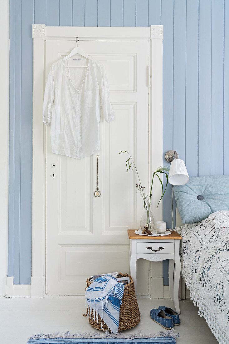 Weiße Bluse hängt an weißer Tür, davor Nachttisch neben Doppelbett im Schlafzimmer mit hellblauer Wand