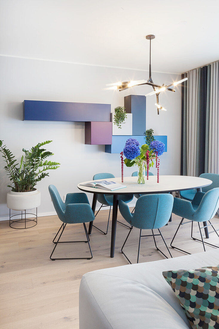 Ovaler Esstisch mit blauen Stühlen in offenem Wohnraum, modulare Regale an der Wand