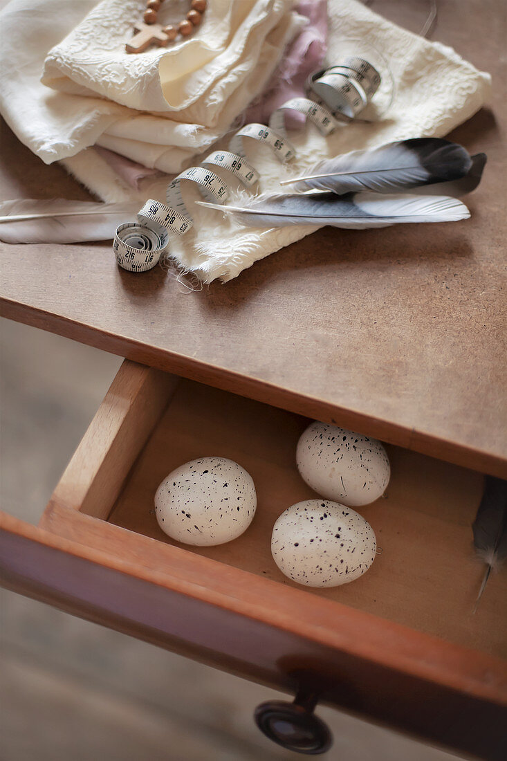 Weißes Tuch mit Federn und auf Tisch, gesprenkelte Eier in geöffneter Schublade