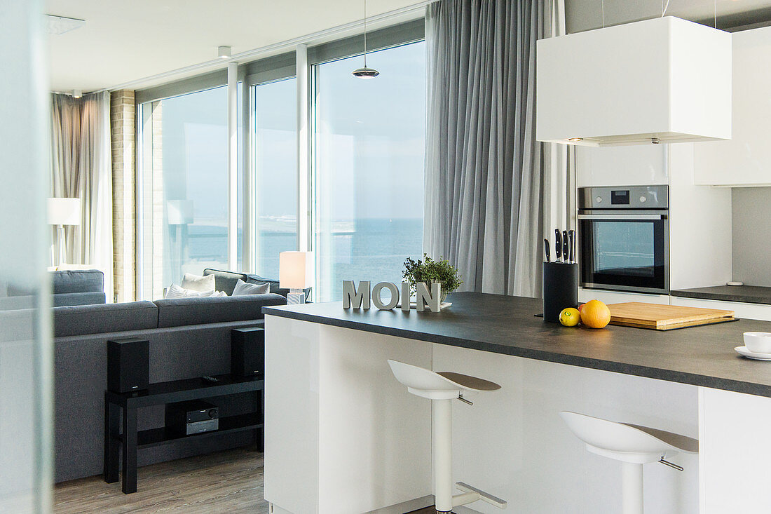 Raumhohe Fensterfronten mit Blick aufs Meer daneben offene Küche mit Küchentheke und Barhockern