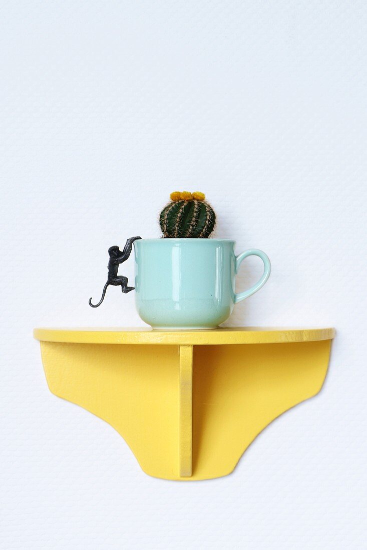 Mit Kaktus bepflanzte Tasse auf einer gelben Wandkonsole