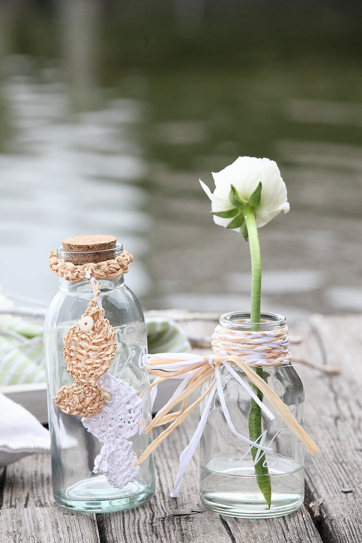 Ranunkel in Glasflasche mit Kranz aus geflochtenen Bastresten und Flasche mit gestrickten Fischen aus Bast